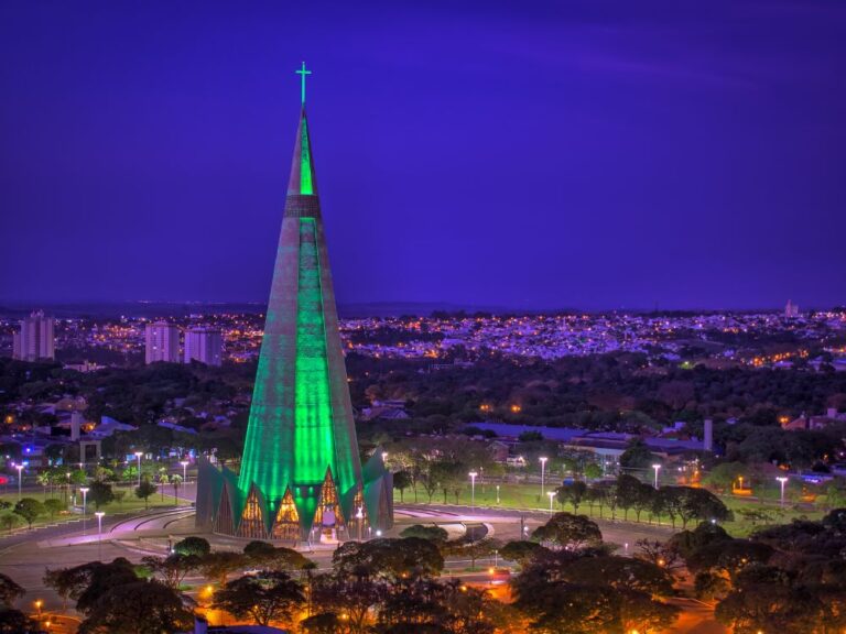 Vista noturna da Catedral de Maringá, um imponente edifício cônico, iluminado em verde vibrante. A estrutura domina o horizonte da cidade, cercada por árvores e luzes urbanas.