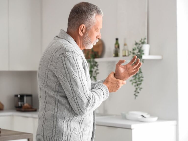 Homem idoso em ambiente doméstico segurando o punho com expressão de desconforto, indicando possível dor nas articulações das mãos ou fragilidade física.