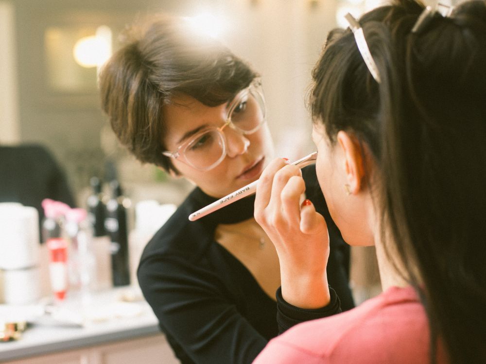 Maquiadora aplica maquiagem nos olhos de uma cliente com um pincel em estúdio de beleza, em close, com iluminação suave.