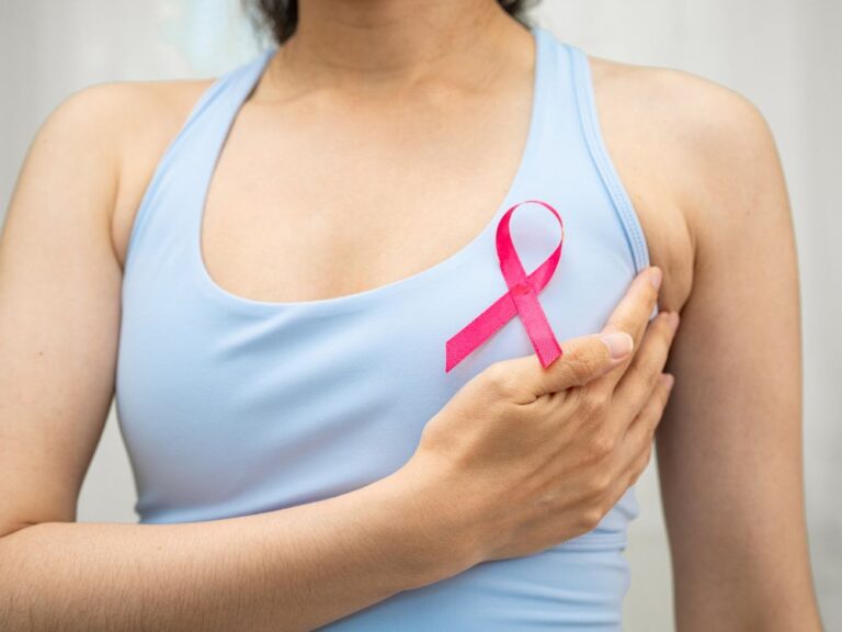 Mulher com laço rosa no sutiã esportivo tocando a região da mama, imagem simbólica de conscientização sobre saúde da mulher e câncer de mama.