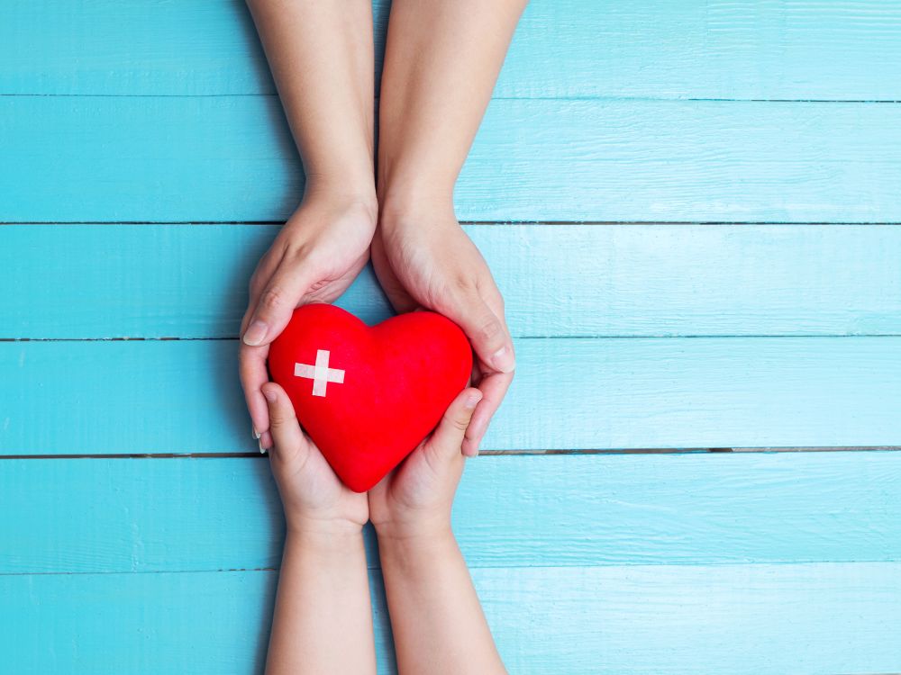 Mãos de duas pessoas segurando um coração vermelho com símbolo de saúde sobre fundo azul, representando cuidado, proteção e bem-estar compartilhado.